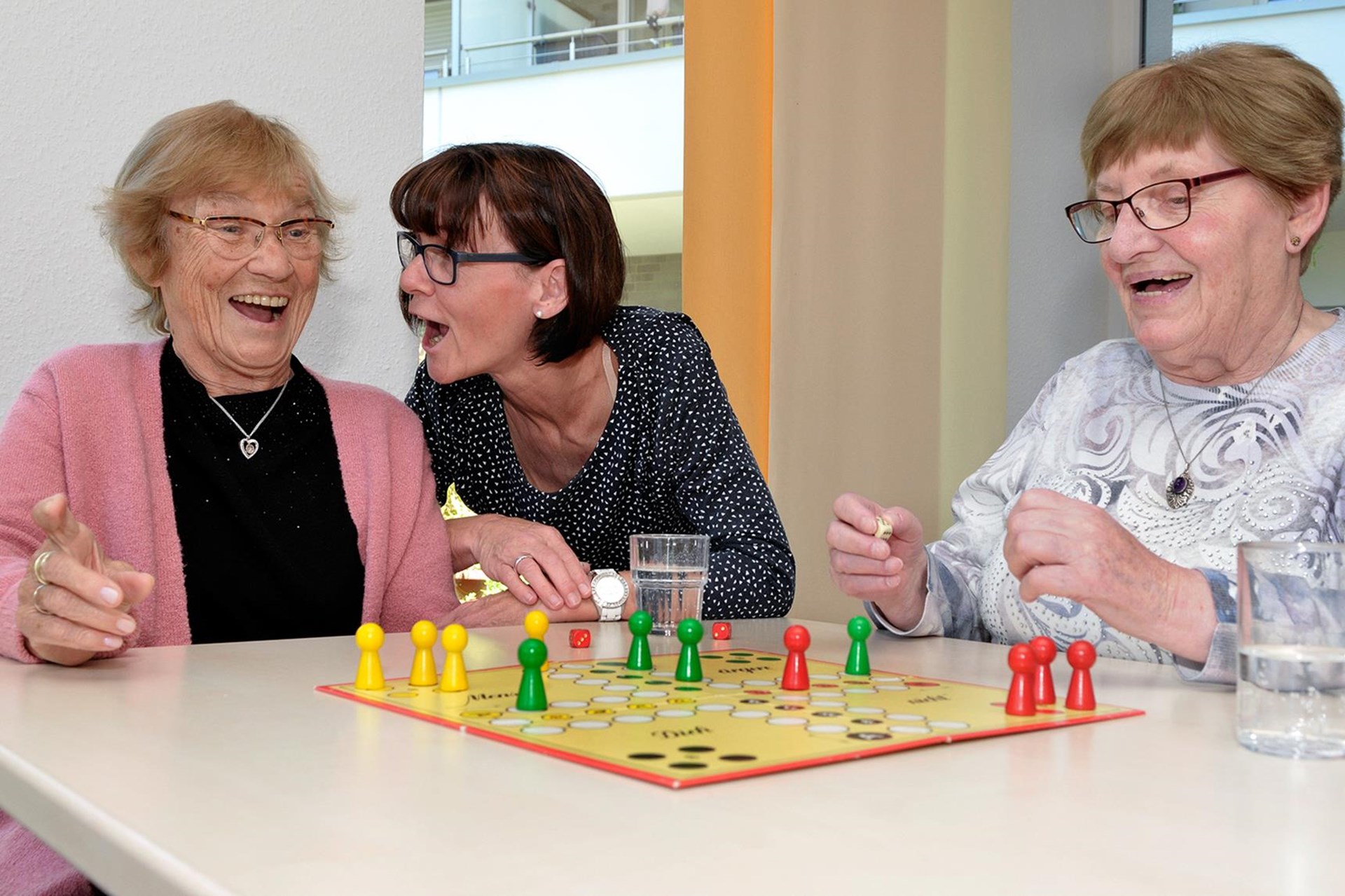 Seniorinnen spielen Mensch-ärgere-dich-nicht, eine Mitarbeiterin feuert sie an